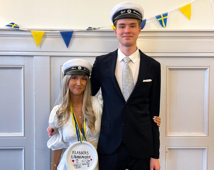 Moa Larssen och Egil Bergström på Thoren Business School Gävle firar studenten. Moa är klädd i vit klänning och studentmössa, Egil har kostym, även han med studentmössa. Bakgrunden pryds av svenska flaggor och vimplar.